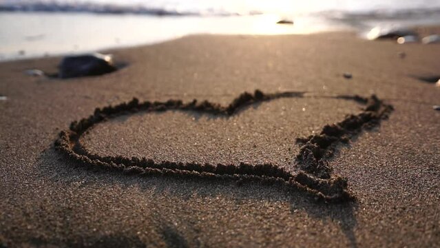 Heart symbol drawn on beach sand at beautiful sunset, washed away by sea waves. Hand drawn heart on beach sand over sunset or sunrise sky beautiful light sea nature landscape. Close-up