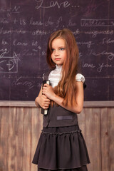 Kid schoolgirl in school uniform holding book at blackboard in classroom, pensive looking. Portrait of cute first grader girl with book indoors. School learning, education concept. Copy ad text space