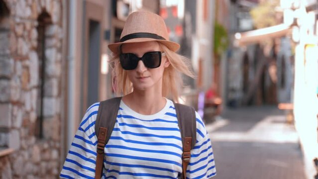 Portrait of caucasian woman, tourist with backpack in straw hat and sunglasses, walking along empty street in old tourist part of town, resting. Sightseeing in popular tourist destination.