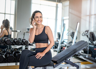 Fototapeta premium Cheerful sporty woman using phone after fitness workout in gym. Young smiling woman athlete using cell phone at gym.Beautiful fit girl with smartphone at fitness centre.