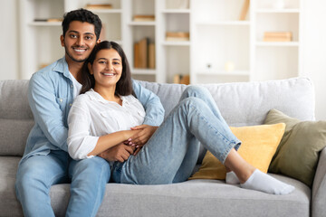 Indian young couple relaxed on sofa, husband hugging wife, sharing a happy moment