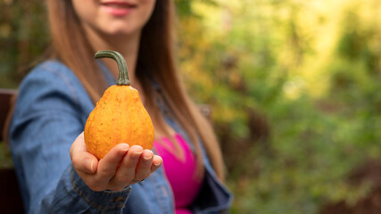 Beautiful young woman with a smile holding a small decorative pumpkin with orange color in nature in the autumn season. Thanksgiving day, fall fruit harvest concept. Close-up.