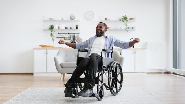 Joyous bearded man in wheelchair exercising stretching arms while following workout program at home. Positive african adult in cozy outfit participating in physical activity in apartment.