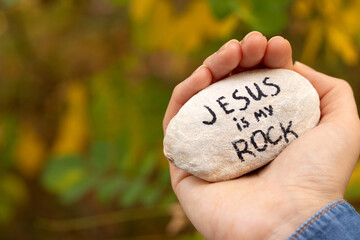 Hand holding a stone with handwriting "Jesus is My Rock" in nature. Close up. Inspiring Christian text verse, solid foundation, strength, and hope in God. Biblical concept.