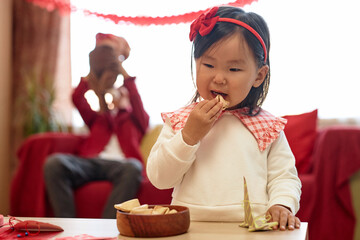 Portrait of cute Asian baby girl eating fortune cookie at home with red decorations for Chinese New...