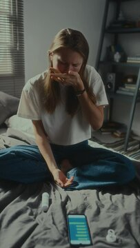 Vertical shot of depressed girl taking handful of pills, lying on bed beside smartphone and crying while suffering from being cyberbullying victim
