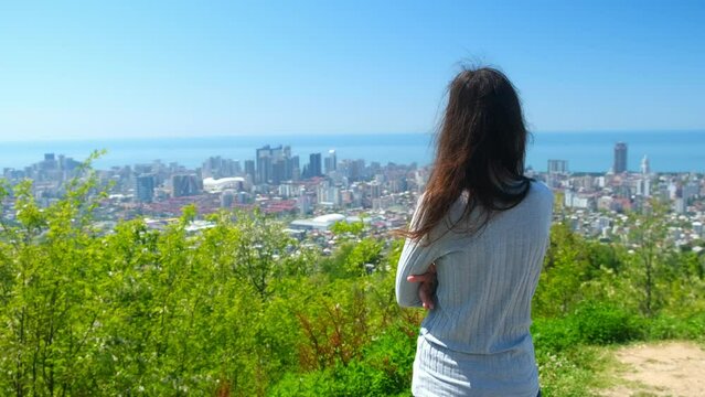 Woman tourisy admiring cityscape of Batumi city from mountain in sunny day. Coastal city in Georgia. Happy smiling female looking at sea, back view. Skyscrapers and slums contrast of living standards.
