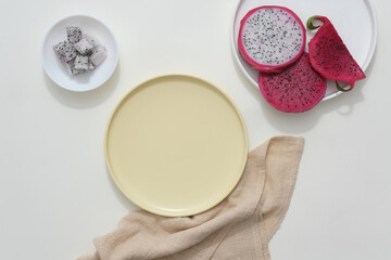 Several plates of different sizes, fresh dragon fruit, and a beige cloth are arranged on a white background. Scene for advertising products related to dragon fruit, with empty space for design.