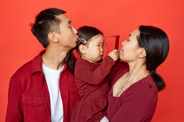 Waist up portrait of modern young Chinese family with baby boy standing against red background together kissing child