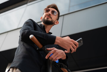 A young hipster man with electric scooter standing in front of modern business building on city street and using a phone.