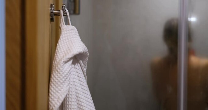 White robe on hook and naked woman taking relaxing shower in morning or evening. Concept of body hygiene and services of spa centers and hotels