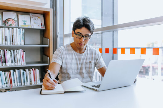 Man with glasses writing in notebook while using laptop in the library