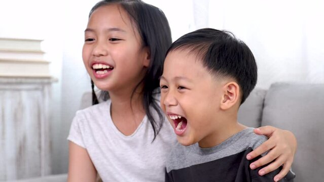 Overjoyed laughing out loud little sibling children watching movies or games together sitting on the floor, happy emotional Asian boy and girl kids have fun bonding relationship stay at home weekend