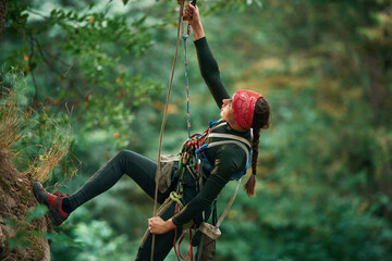 In sportive clothes, high up on the rope. Woman is doing climbing in the forest by the use of safety equipment - obrazy, fototapety, plakaty