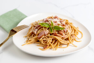Spaghetti carbonara with cheese and basil on white plate with golden fork on white marble background,