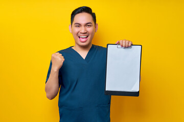 Excited professional young Asian male doctor or nurse wearing a blue uniform holding clipboard with blank paper and celebrating success isolated on yellow background. Healthcare medicine concept - obrazy, fototapety, plakaty