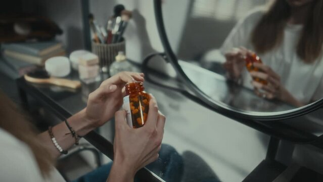 Tilt up shot of stressed girl taking pills from bottle and looking at reflection in mirror while attempting to commit suicide at home