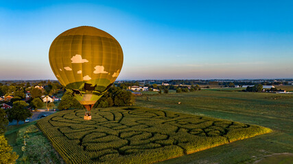 An Aerial View of a Golden Hot Air Balloon, Just Launched and Floating Across a Field With a Corn Maze, on a Sunny Summer Morning