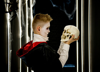 little boy as halloween vampire in red and black hood and black teeth holding  skull in his hands