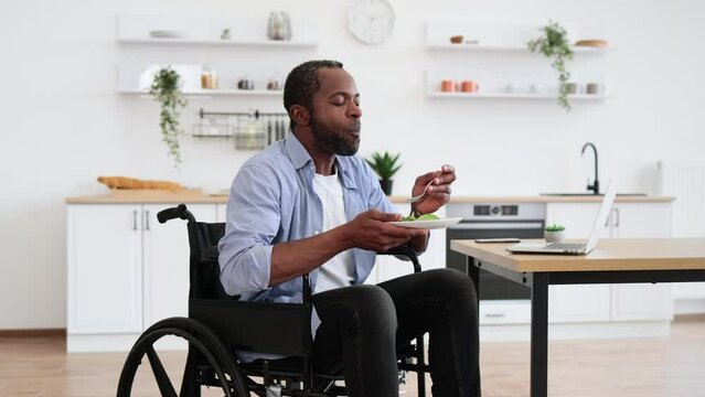 African bearded male in wheelchair using computer during mealtime in kitchen interior. Business manager increasing energy with vegetarian salad on plate while staying in remote office.