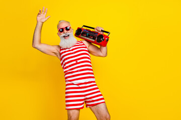 Photo of smiling happy aged man with long beard dressed striped red swimsuit dancing hold boombox isolated on yellow color background