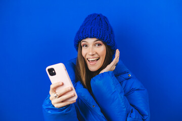 Shocked brunette woman in blue puffer jacket hold mobile phone isolated on blue background studio....