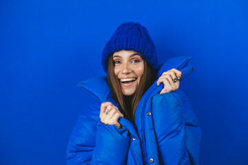 Stylish happy woman holding puffer jacket over blue background. Girl wear total blue outfit, hat,...