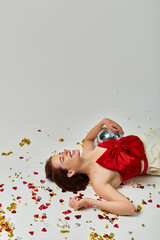 New Year party, excited young woman with disco ball lying on floor near confetti on grey backdrop