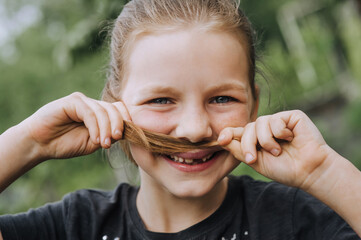 Beautiful red-haired smiling cheerful funny girl, the child holds her hair in her hands, depicting a mustache near her lips. Photography, face close-up portrait.