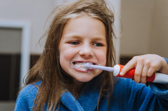 A little girl teenager, a child in a bathrobe cleans her teeth with a toothbrush while looking at herself in the mirror in the morning. Photography, childhood concept.