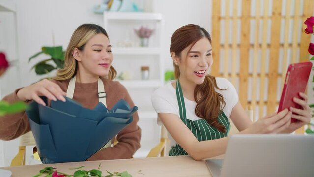Two young Asian women owner floral store holding rose flowers on hands showing to client through video call on tablet for taking order. Small business concept 