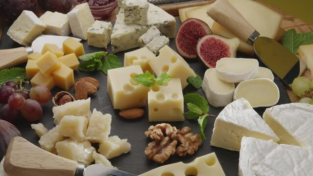Variety of sliced cheeses with fruits, mint, nuts and cheese cutting knives slowly rotate in the frame. A wonderful cheese background for your projects.