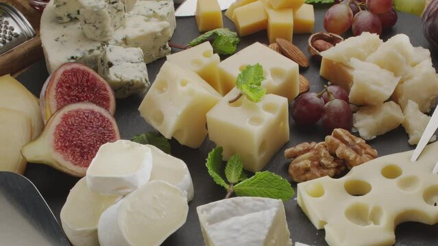 Variety of sliced cheeses with fruits, mint, nuts and cheese cutting knives slowly rotate in the frame. A wonderful cheese background for your projects.