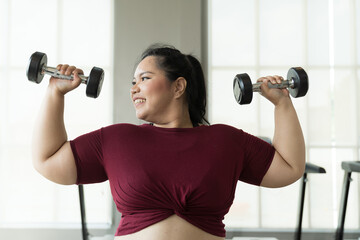 Asian young chubby size woman lifting dumbbells weight in fitness club. Young overweight woman...