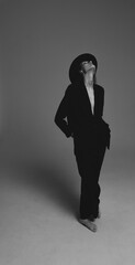 Black and white portrait. A beautiful young girl in black trousers, a black hat and jacket on a naked body stands on a white background