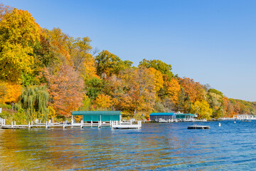 Obraz premium Brilliant autumn trees along a lake with white piers and blue-green boat covers. and a blue sky.