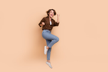 Full length photo of pretty excited girl dressed brown shirt jumping high rising fists empty space...