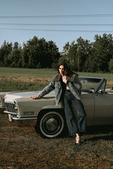 A beautiful woman stands next to a convertible in a field. Dressed in jeans and a jacket with fur. Beautiful and stylish look