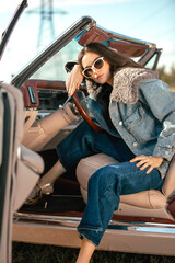 A beautiful woman sits on a convertible in a field wearing sunglasses. Dressed in jeans and a fur...
