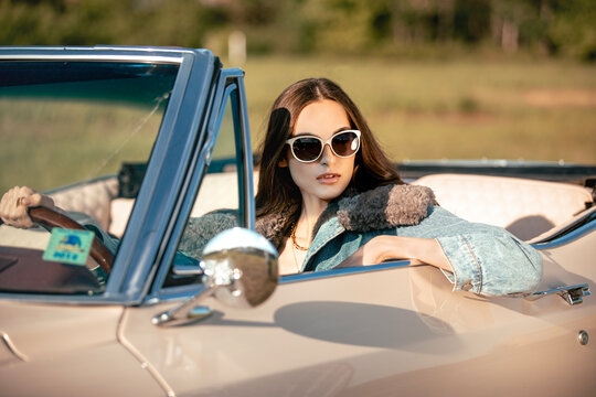 A beautiful woman sits on a convertible in a field wearing sunglasses. Dressed in jeans and a fur jacket. Beautiful and stylish look