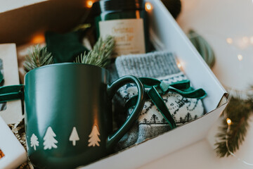 Close-up of Christmas gift box. Cozy mug for coffee, warm gray socks, aroma candle in jar....