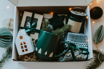 Christmas care package gift box. Paper box with cozy mug, candles, socks, candies and white ceramic house. Christmas and Holiday Gift Ideas - 667750844