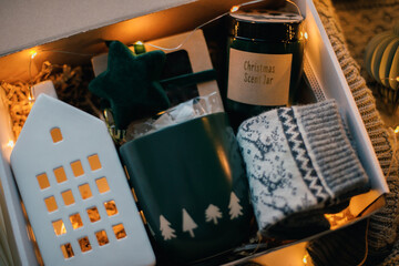 Christmas and Holiday Gift Ideas. Winter present gift box. Cozy mug for coffee, warm gray socks, aroma candle in jar, white ceramic house. - 667751081