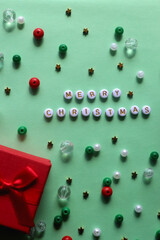 Small red and white Christmas presents, letter beads spelling Merry Christmas and various colorful beads on green background. Top view.