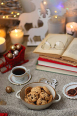Bowl of cookies, cup of tea, dry oranges, pine cones, books, reading glasses, small presents, various Christmas decorations and lit candles on the table. Cozy Christmas hygge. Selective focus.