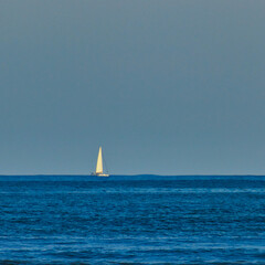 image in square format, of a white sailboat sailing on the horizon in a wavy cobalt blue ocean,...