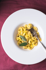 Tortellini with sage leaf is a festive winter and Christmas dish in Northern Italy. Winter Italian cuisine. Burgundy linen natural background. Close-up
