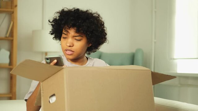 Unhappy angry african woman unpack delivered box receiving damaged shipping mistake problem in postal service. Annoyed confused customer client receiving wrong parcel checking online app on smartphone
