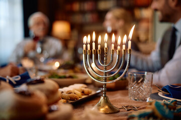 Lit menorah on dining table with Jewish family celebrating Hanukkah in background.