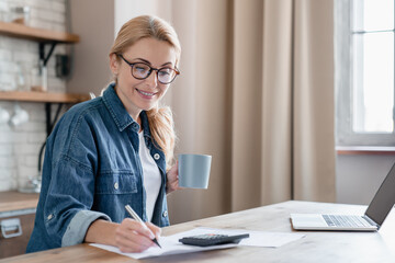 Happy smart mature woman financial advisor taking notes, concentrated on home bills calculations,...